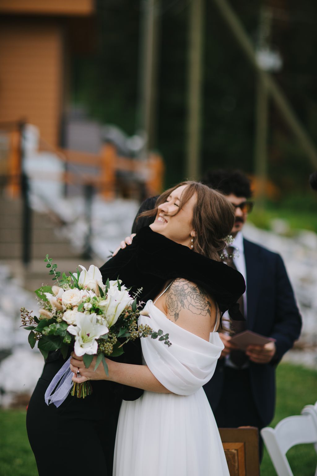 Bride in white dress with black velvet shawl holds white floral bouquet, smiling with eyes closed during outdoor ceremony