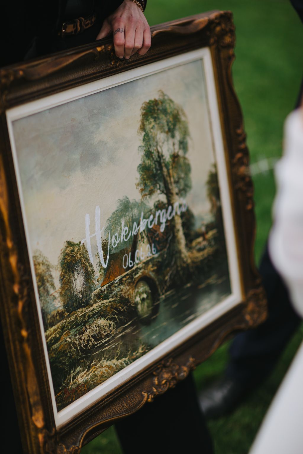 Hand resting on ornate gold frame holding landscape painting with green foliage and handwritten text overlay