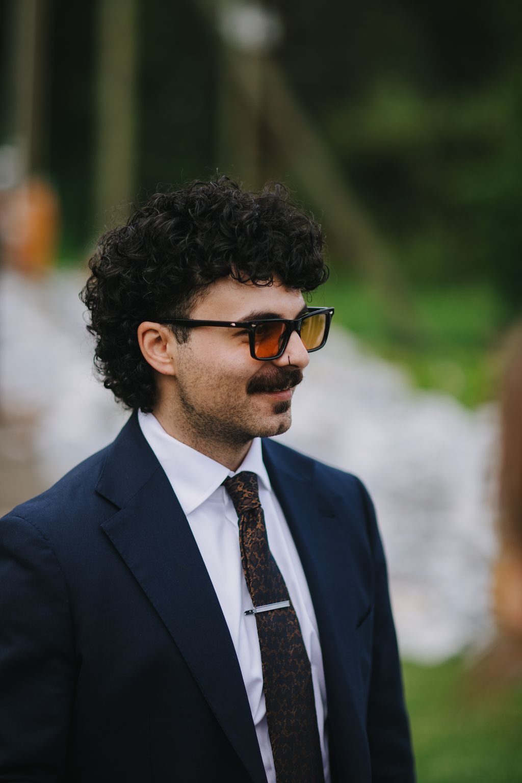 Groom in navy suit and sunglasses with styled dark curly hair, outdoor portrait with blurred green background