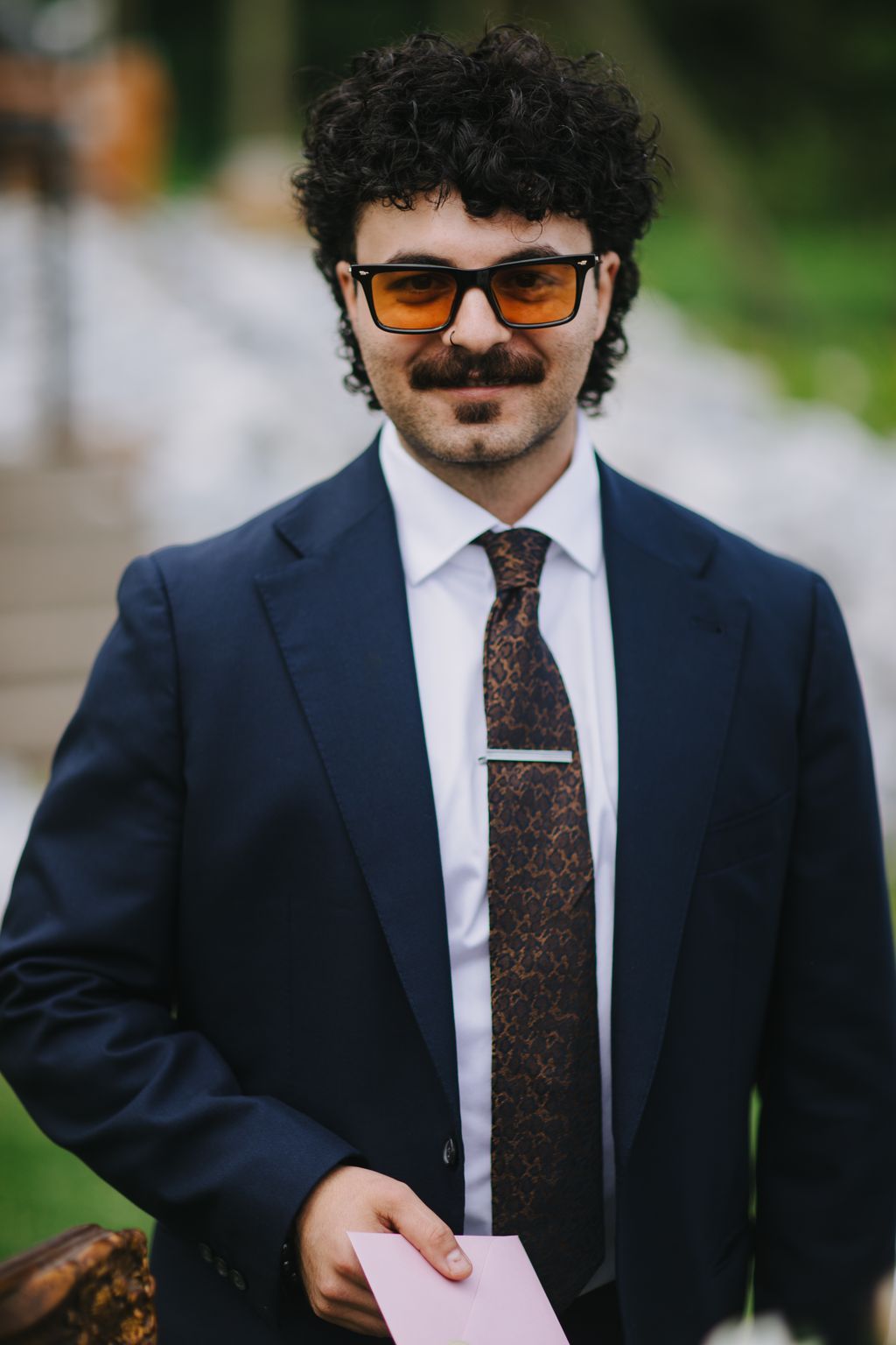 Groom in navy suit with patterned tie and orange-tinted sunglasses holding pink card outdoors