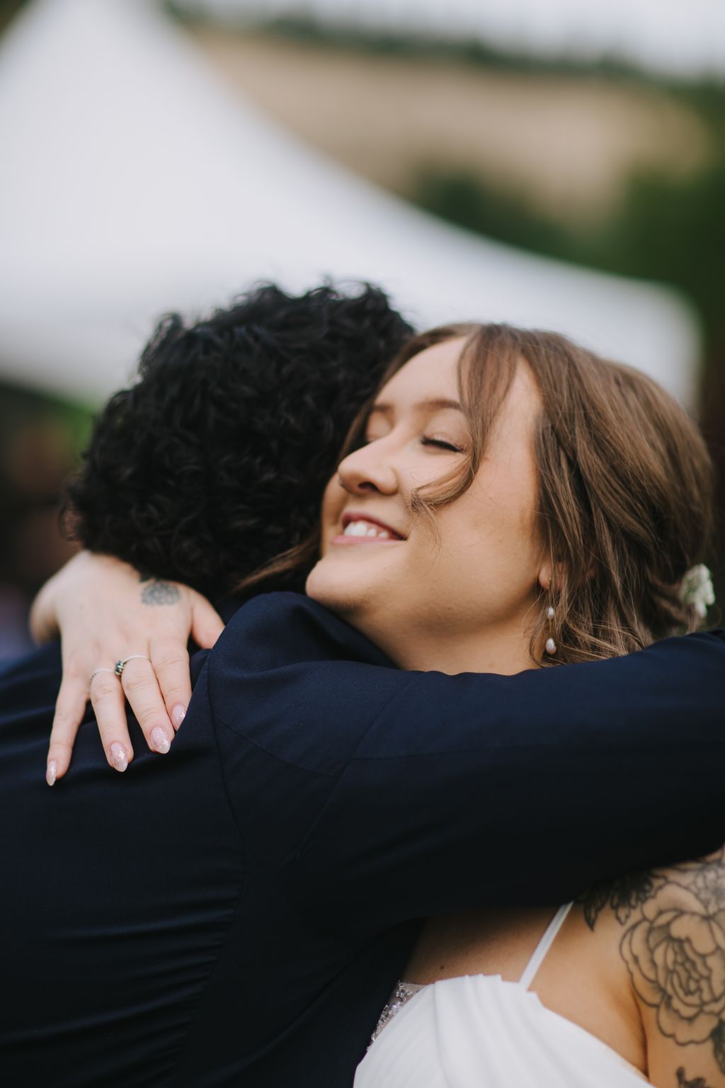 Bride smiling with eyes closed while embracing groom in dark suit against blurred outdoor background