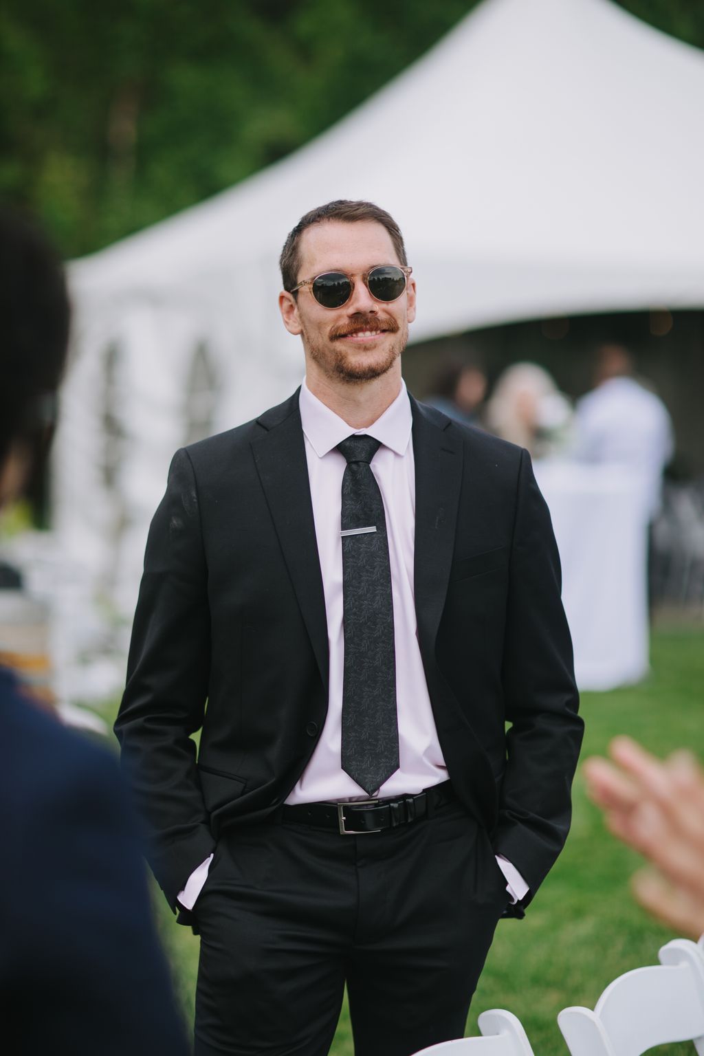 Groom in black suit, white shirt, and dark tie wearing sunglasses smiles at outdoor wedding event