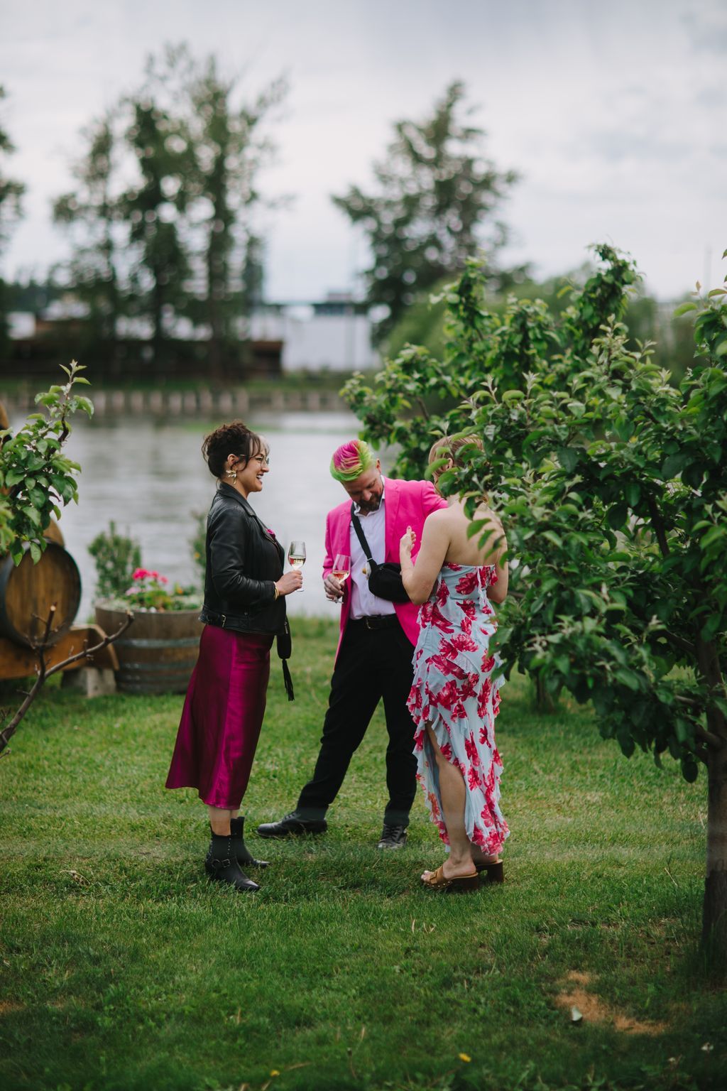 Three guests in vibrant attire enjoy champagne on a manicured lawn with water views and greenery in soft, overcast light