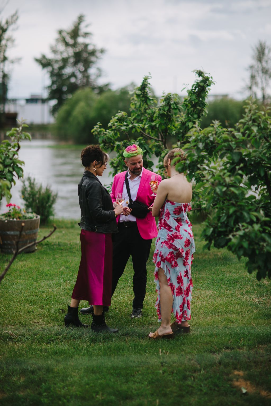 Three guests in vibrant attire toast with champagne glasses on a grassy lawn beside green foliage and water