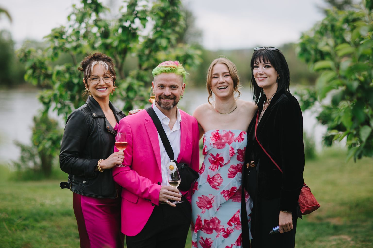 Two smiling people at an outdoor celebration by a water''s edge, one wearing a vibrant pink blazer and the other in a blue floral dress, holding champagne gla…