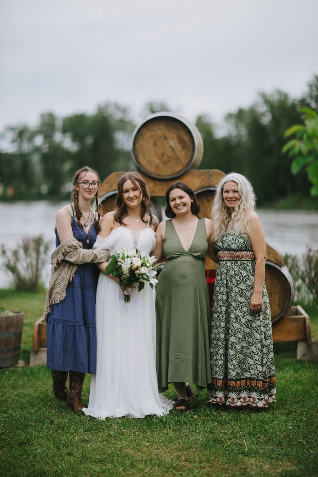 Bride in white dress holding white bouquet poses with three bridesmaids against rustic wooden barrels at outdoor vineyard wedding venue