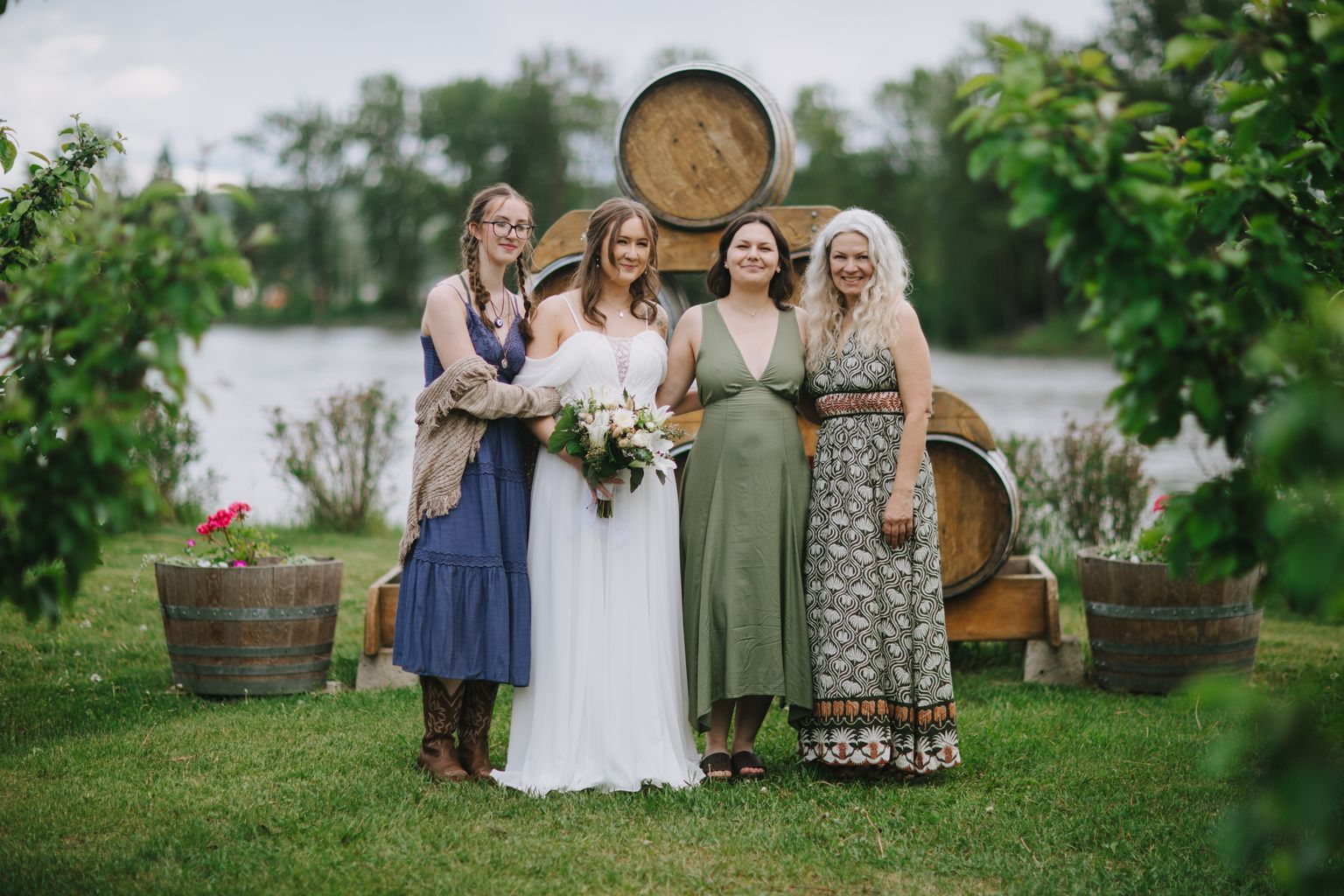 Bride in white dress with bouquet stands with three women in blue, green, and patterned dresses before a wooden barrel at an outdoor venue
