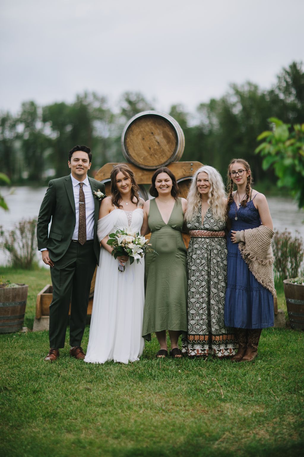 Bride in white gown with bouquet stands with groom and two women beside a wooden barrel at a vineyard, green trees blurred in background