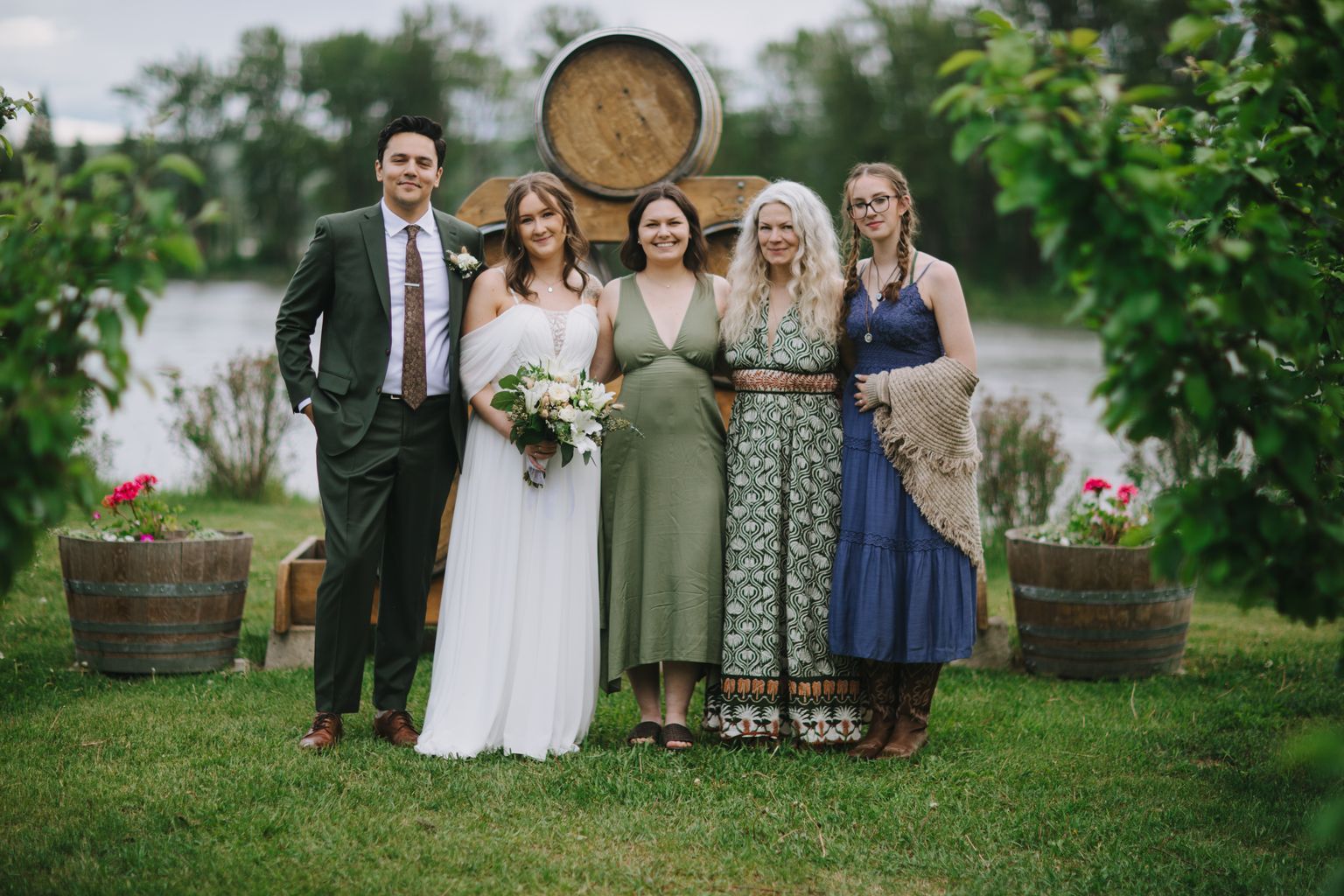Bride in white dress with bouquet poses with three women in varied colored dresses in front of a wooden barrel at an outdoor wedding venue