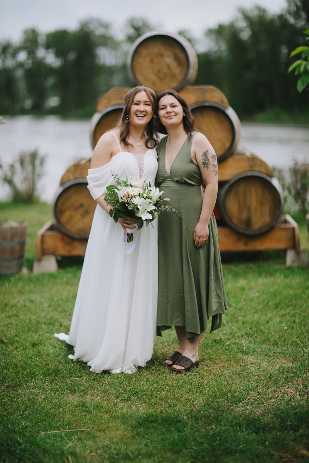 Two women smile at camera in front of wooden barrels, one in white wedding dress holding white bouquet, one in sage green dress