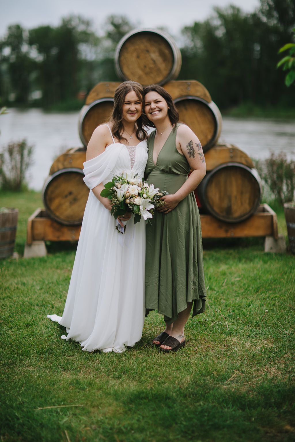 Two women smile at the camera in front of stacked wooden barrels, one wearing a white off-shoulder gown and the other in a sage green dress, holding a white…