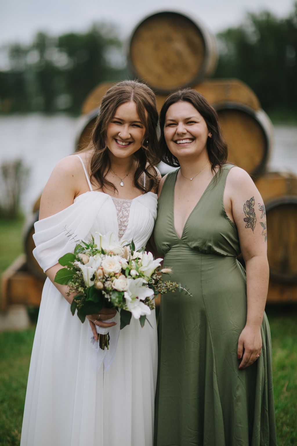 Two smiling women in formal attire holding a white flower bouquet with wooden barrels blurred in background