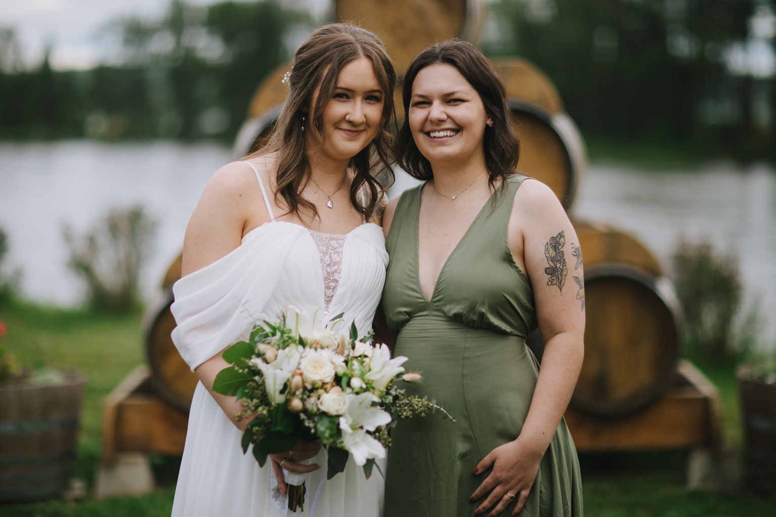 Two smiling women in elegant dresses pose together holding a white floral bouquet outdoors