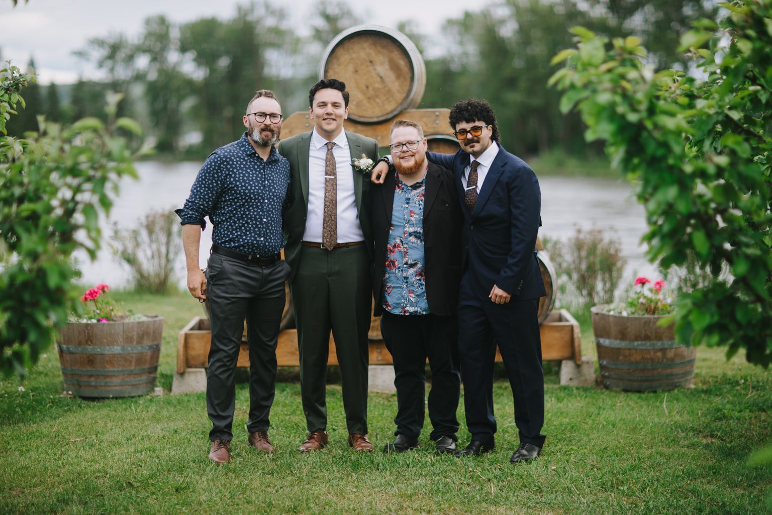 Four groomsmen in suits stand on grass in front of wooden barrels, smiling at the camera in natural outdoor light