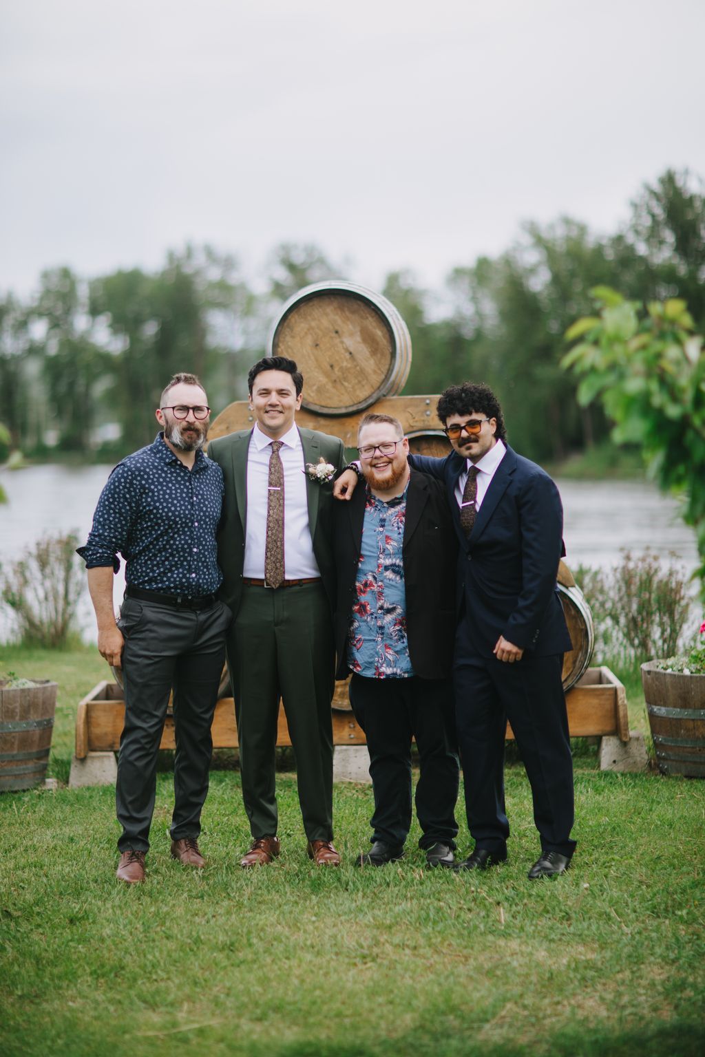 Four men in formal attire stand smiling before wooden barrels and evergreen trees on an overcast day