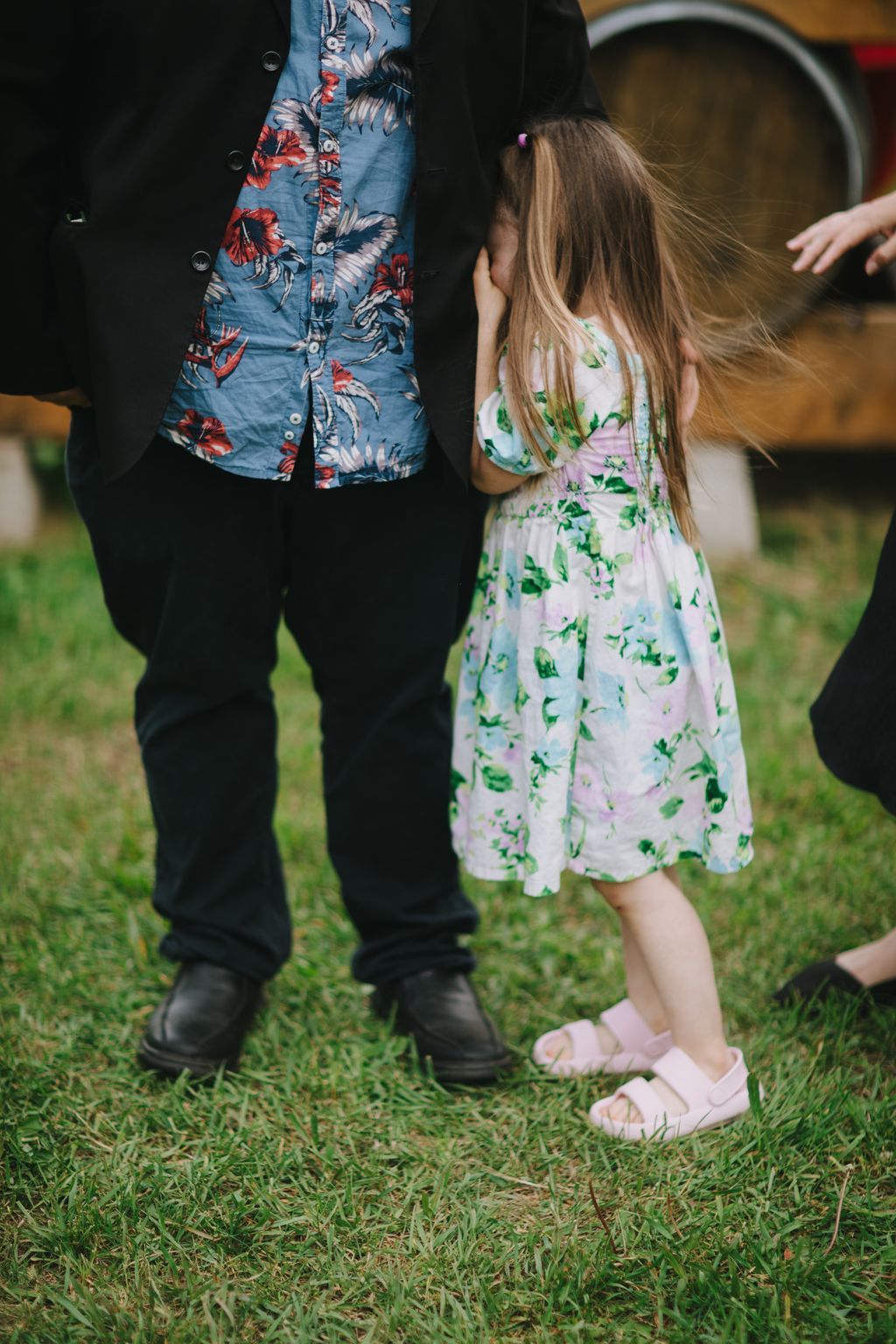 Young flower girl in pink floral dress stands beside groom in blue patterned shirt on grass during outdoor wedding ceremony