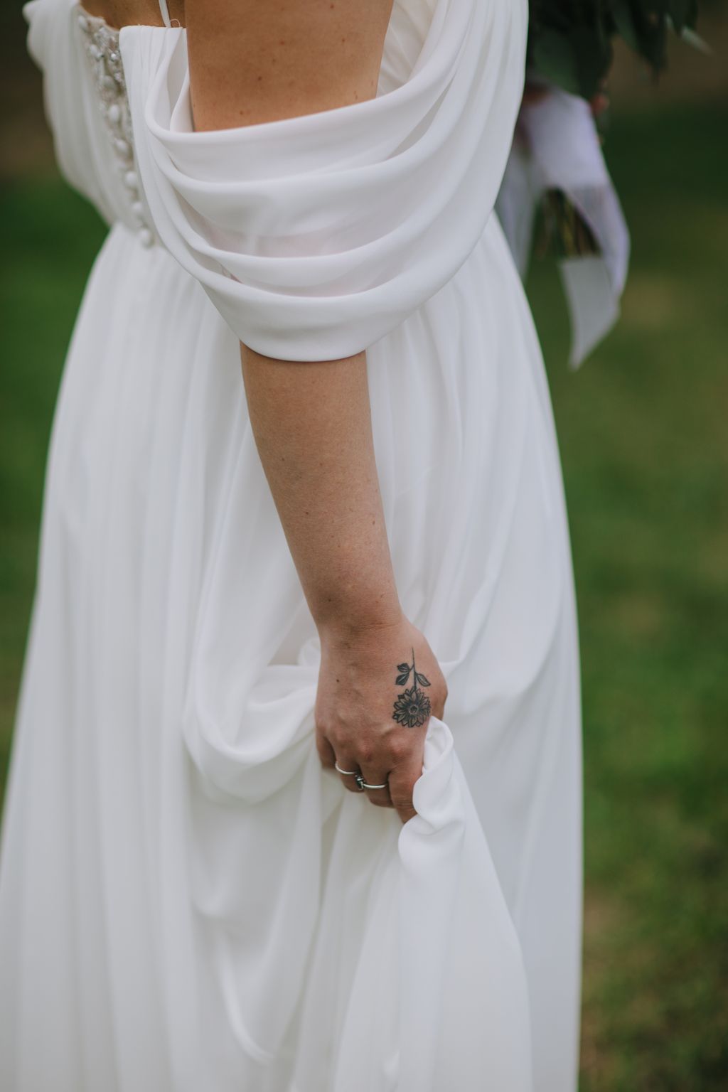 Bride in flowing white dress holds fabric, displaying delicate hand tattoo and simple wedding rings in natural light