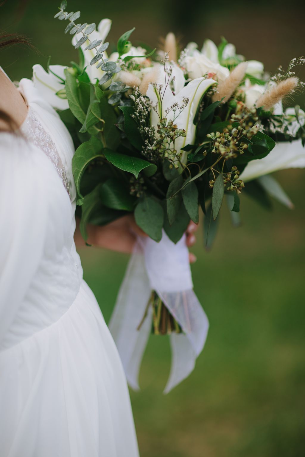 Bride holding romantic white bouquet with eucalyptus, baby''s breath, and delicate greenery tied with white ribbon against soft green background