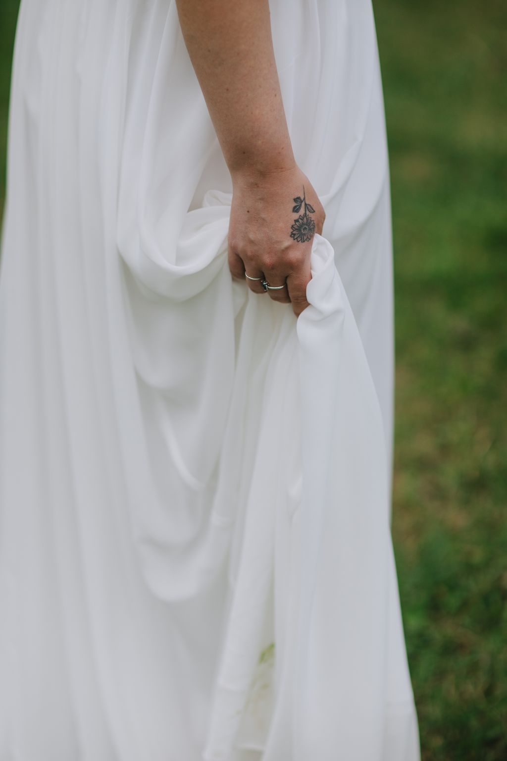 Bride''s hand with sunflower tattoo holding groom''s hand, displaying wedding rings against flowing white dress fabric