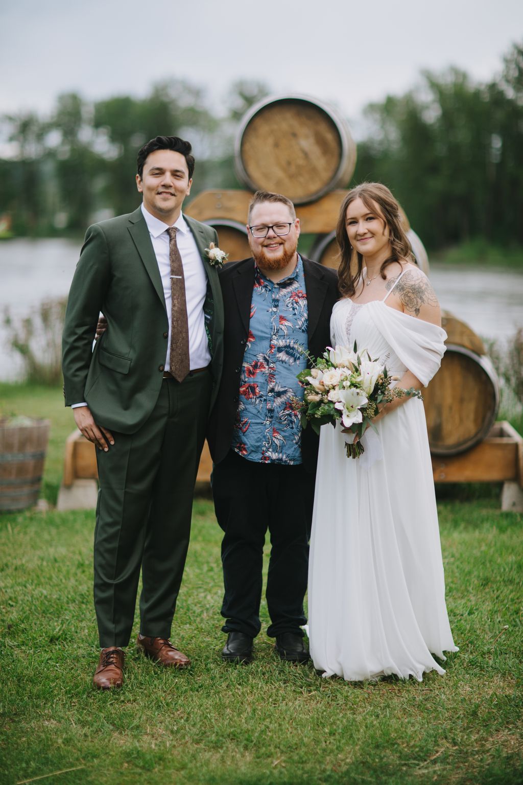 Bride in white dress with bouquet, groom in floral shirt, and groomsman in green jacket smile on grass with wooden barrels behind them