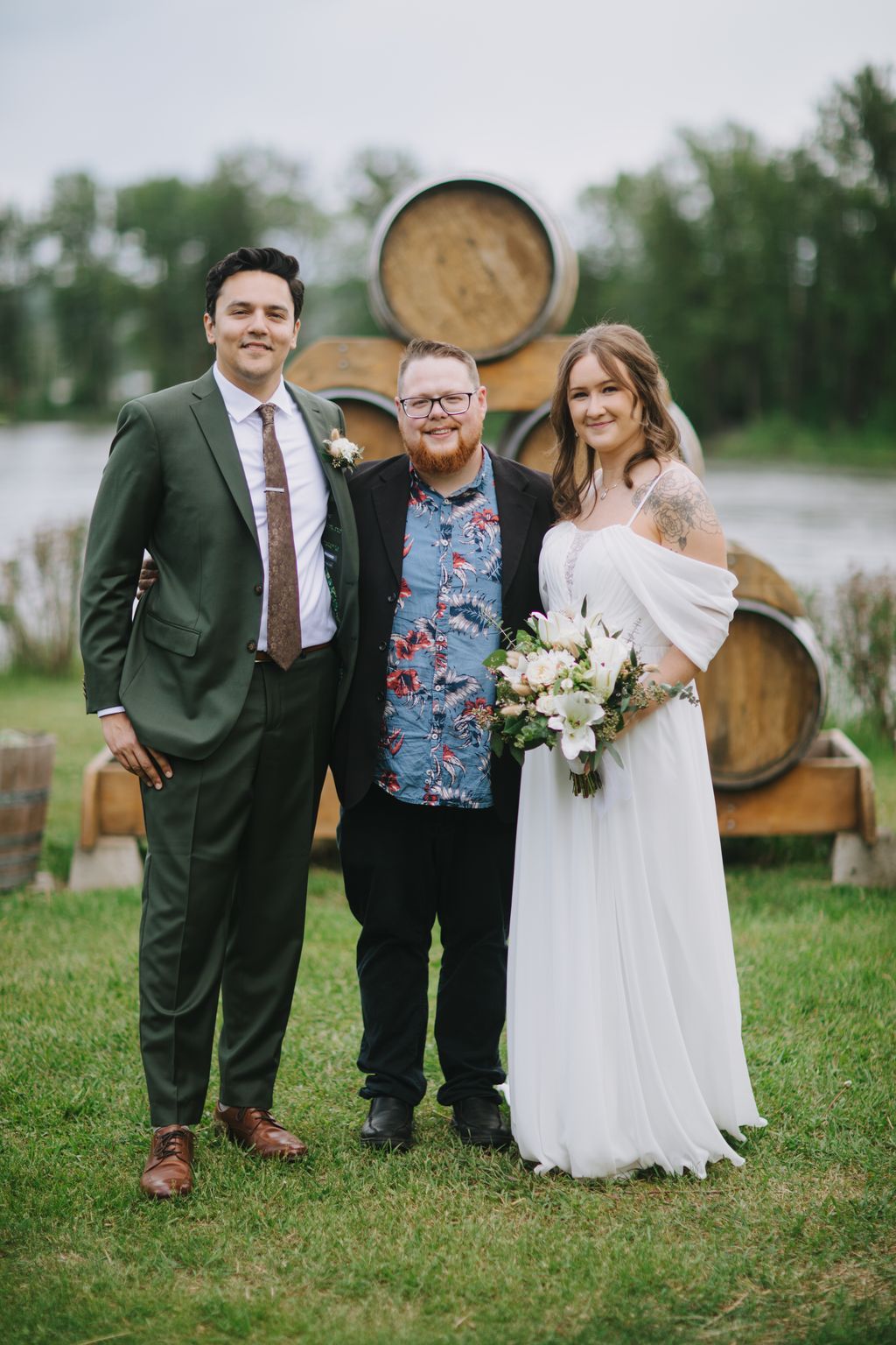 Bride in white dress holding bouquet poses with two groomsmen beside wooden barrels at outdoor vineyard venue