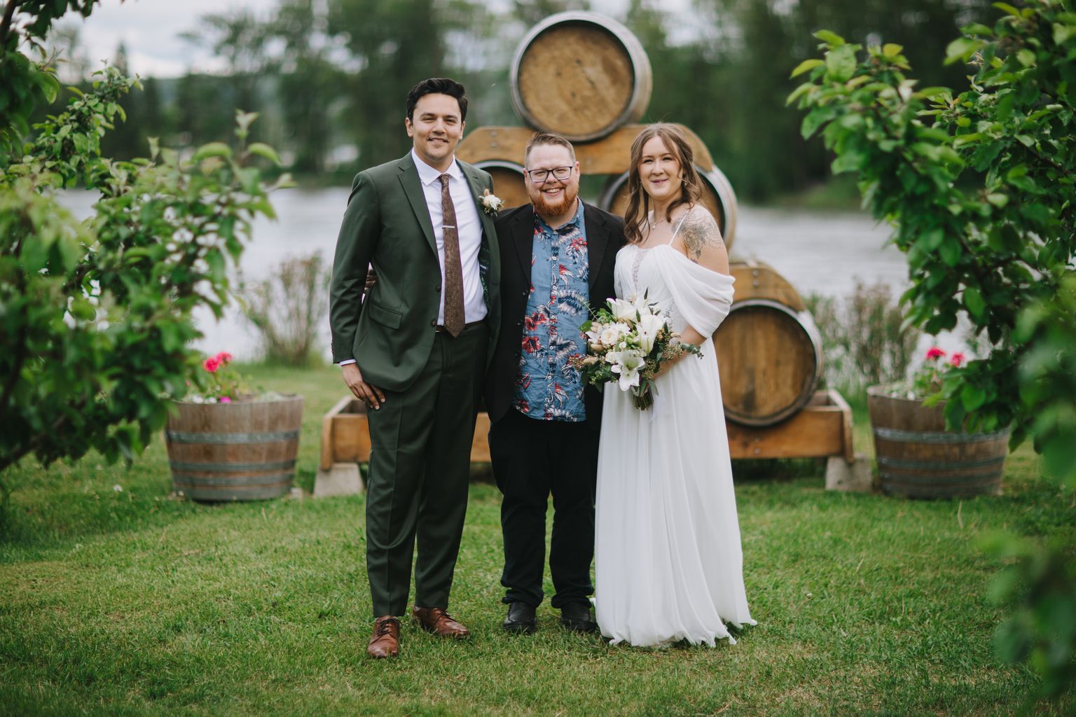 Bride in flowing white dress with groom and officiant standing on grass before wooden barrels at outdoor wedding