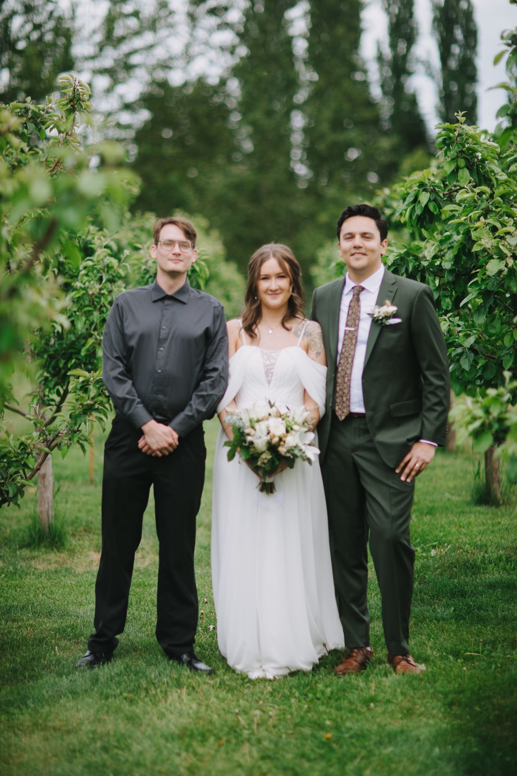 Bride in white dress holding bouquet stands between two groomsmen on a grass lawn with tall trees in soft-focus background