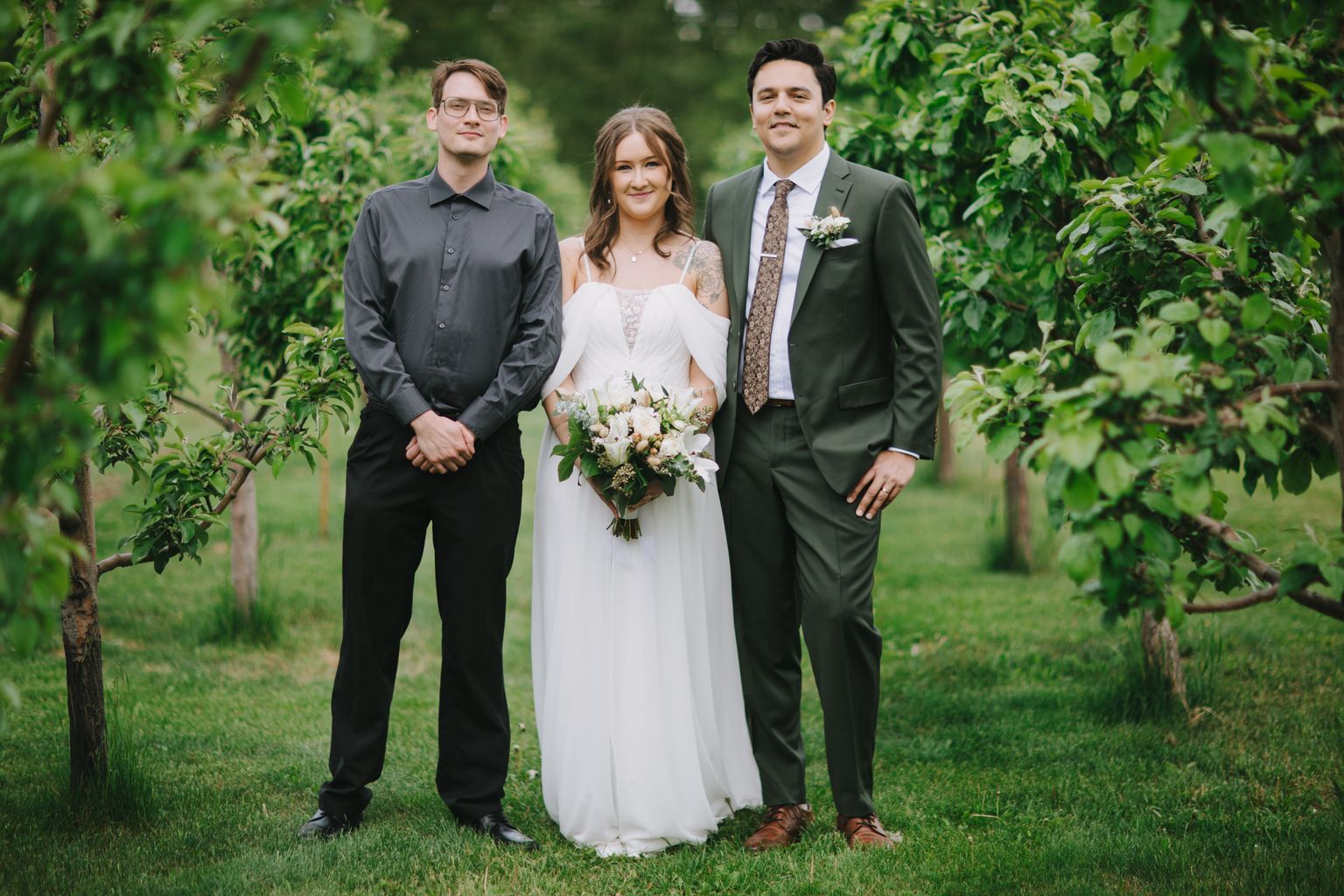 Bride in white cold-shoulder gown holding greenery bouquet stands with groom in sage suit and groomsman outdoors on grass