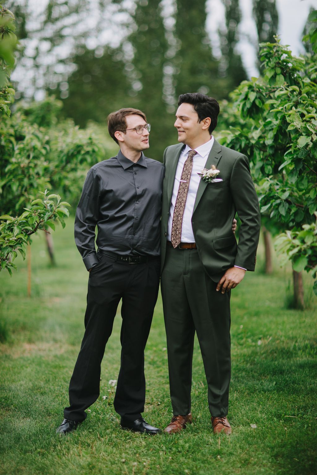 Two grooms smile at each other on a grass lawn with tall green trees and dappled light in the background