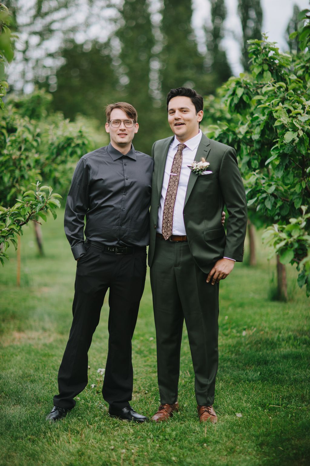 Two men stand on a grass lawn with tall evergreen trees in soft focus behind them, one wearing a dark shirt and glasses, the other in a sage green suit with…