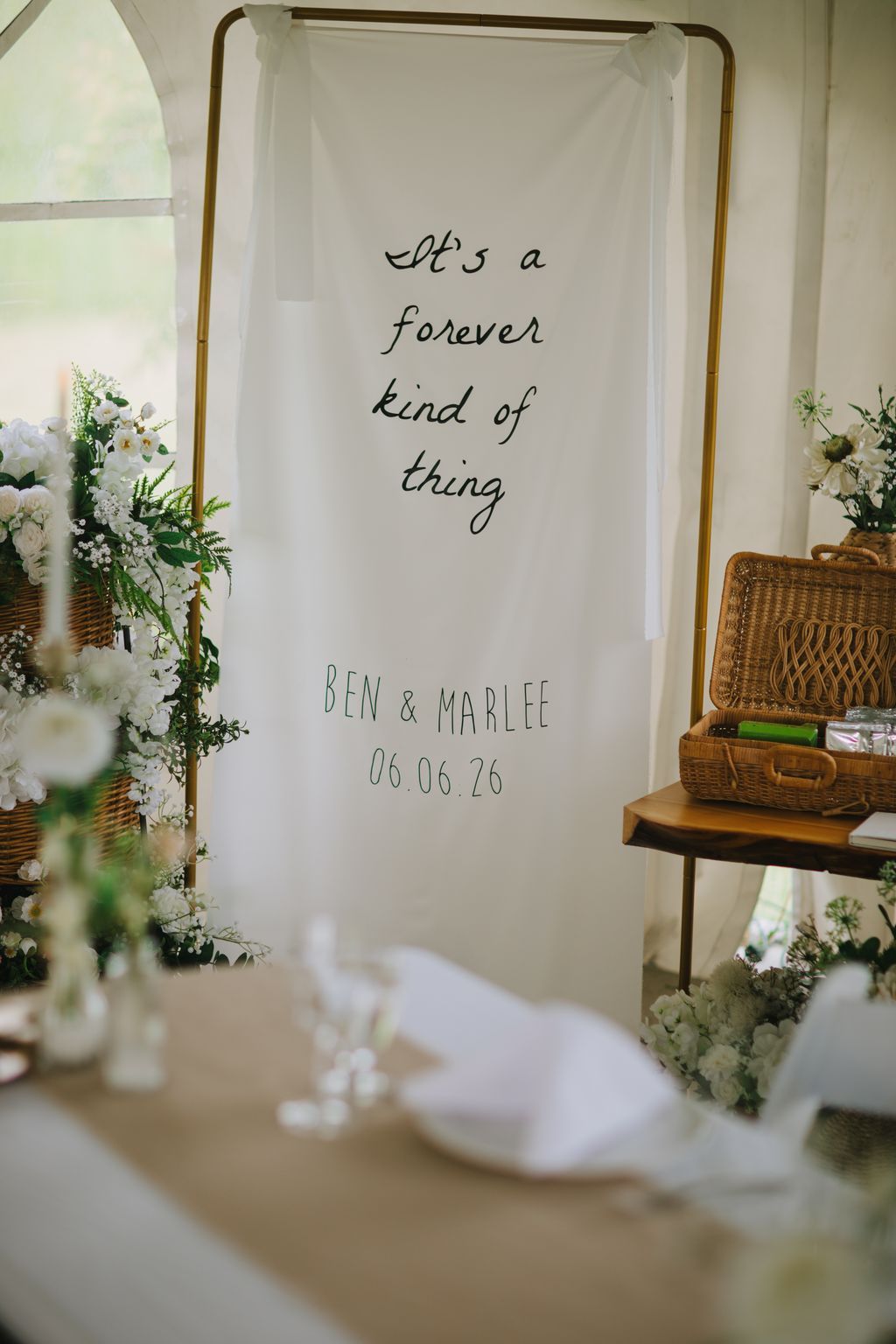 Hand-written veil with "It''s a forever kind of thing" text and "BEN & MARLEE 06.06.26" displayed on wooden rod with greenery accents