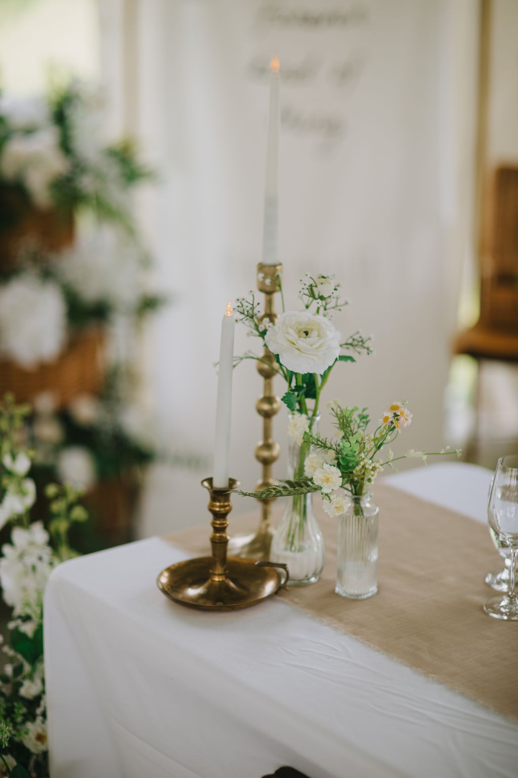 Elegant wedding tablescape with white roses in glass vases beside brass candlesticks holding lit white tapers against a soft, blurred background