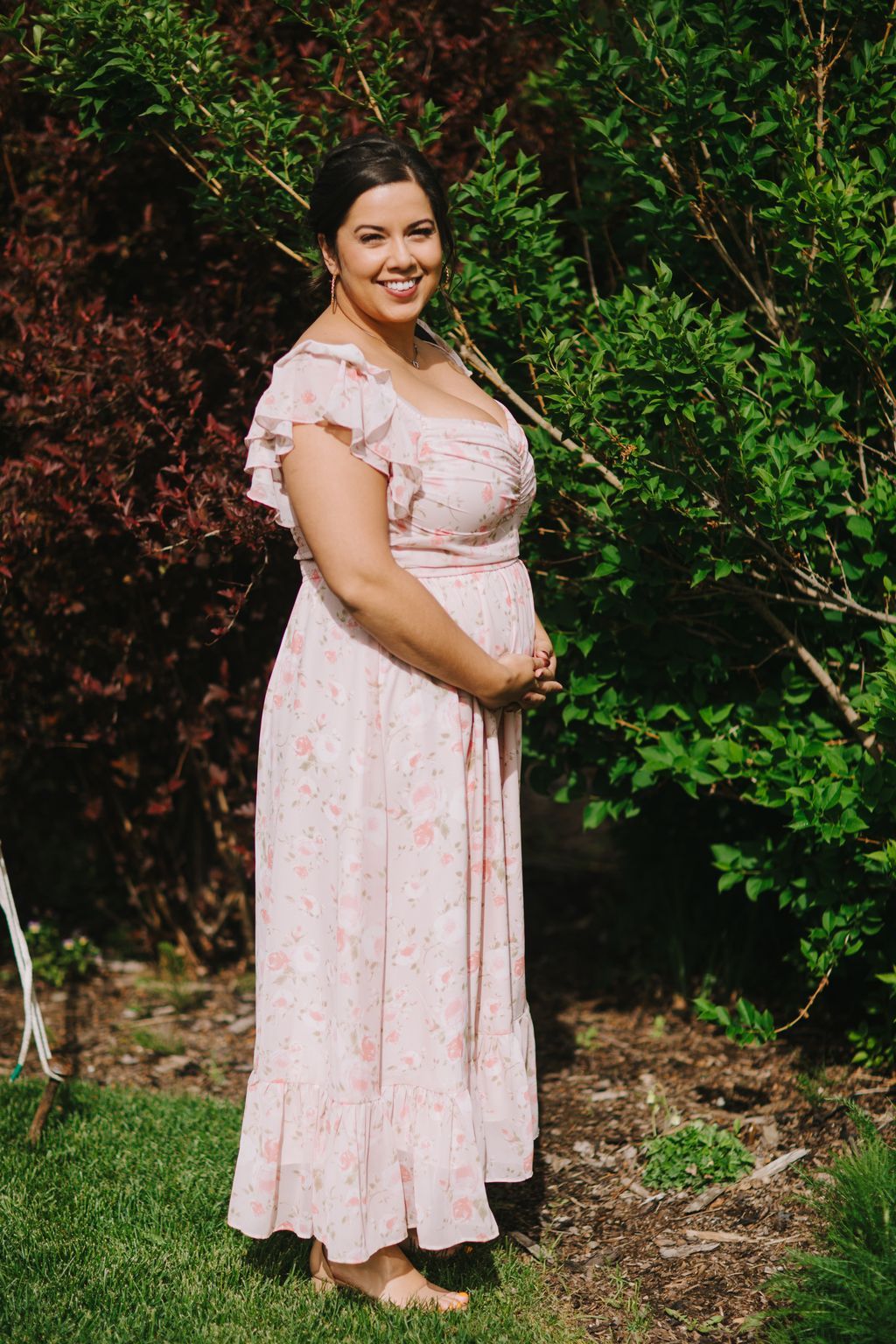 Pregnant woman in pink floral dress smiling at camera against lush green foliage backdrop