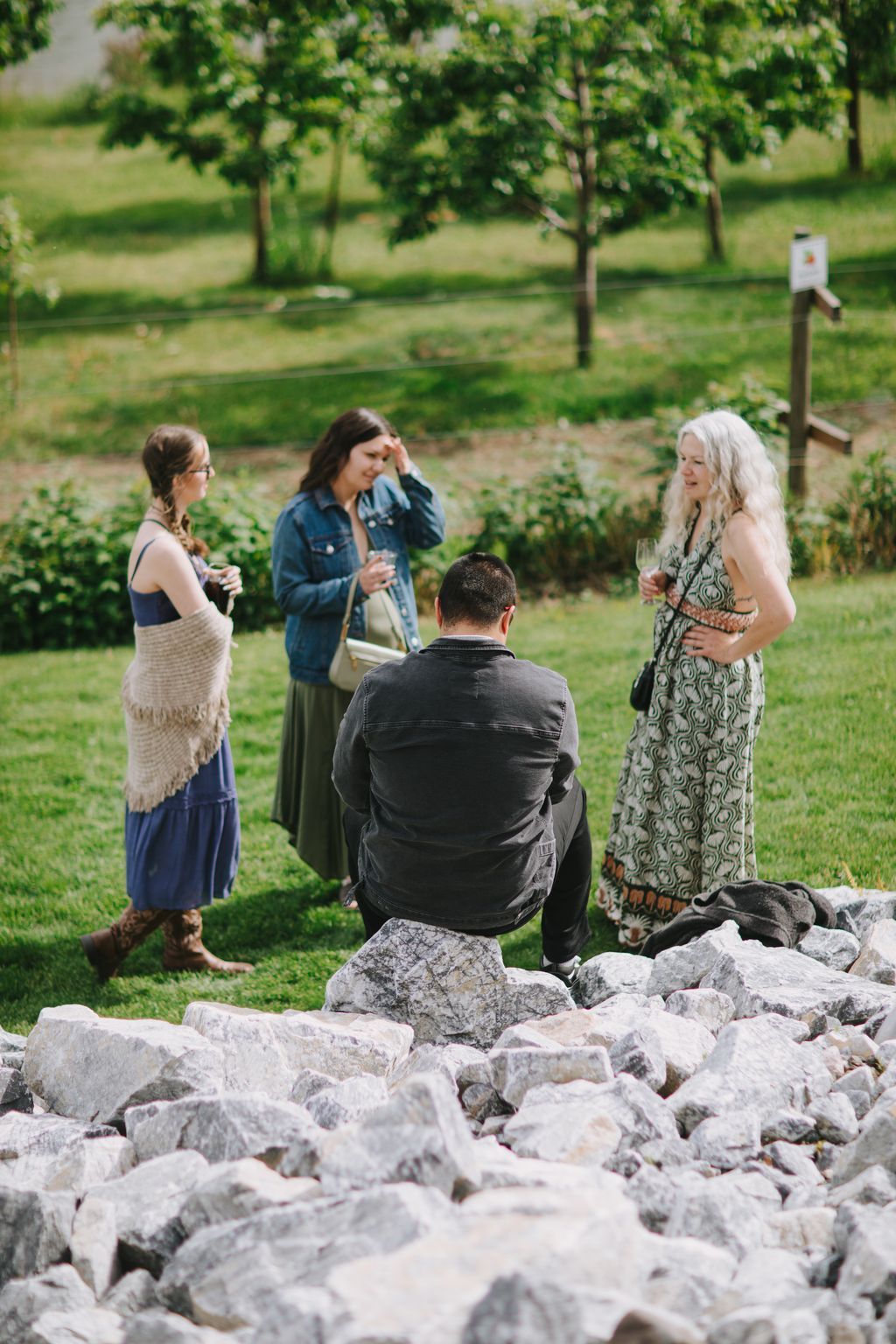 Couple stands together in a lush garden setting with trees and green grass, woman in denim jacket touches her face thoughtfully while holding a glass