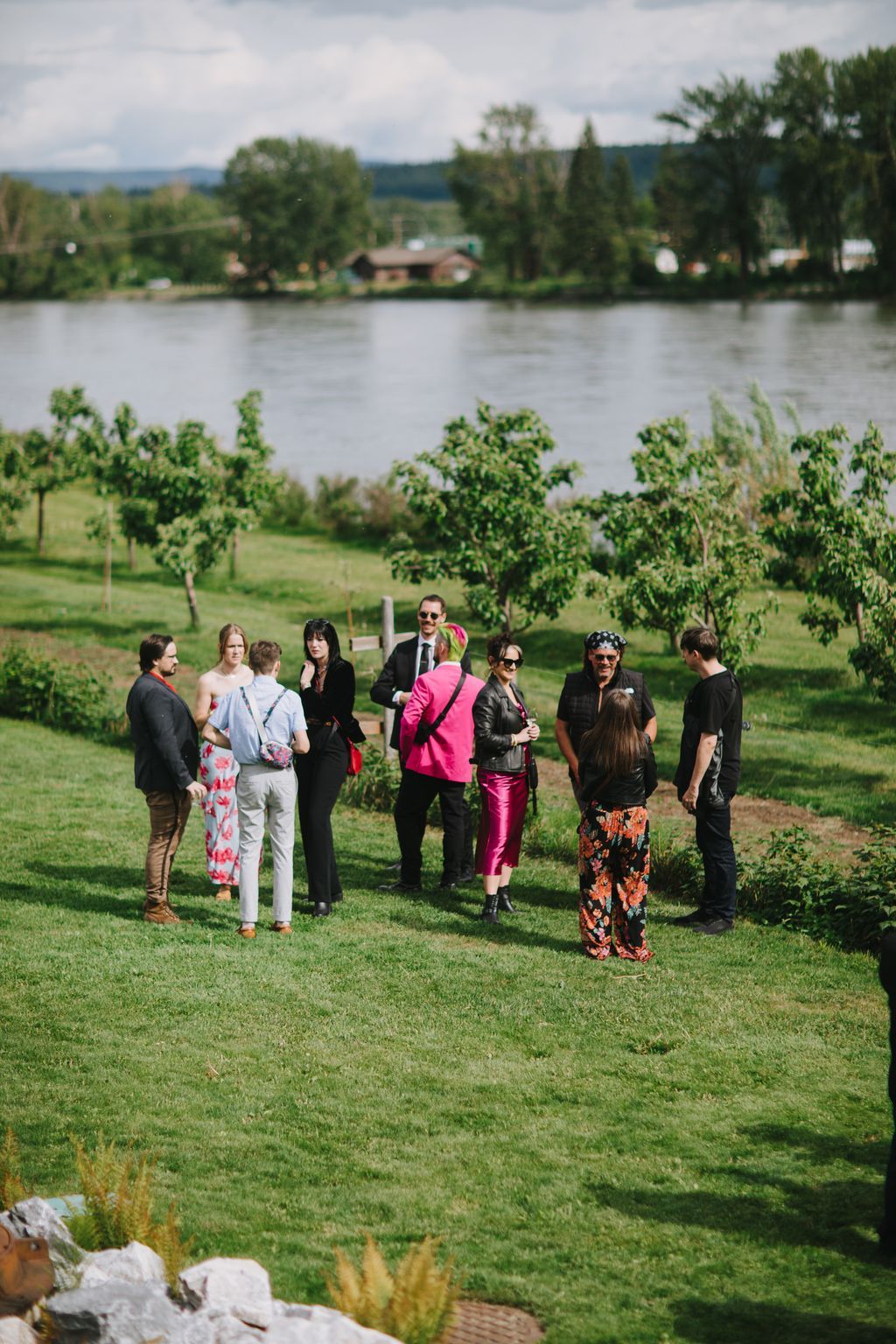 Wedding guests gather on a grassy riverside lawn with trees and a house visible across the water under cloudy skies