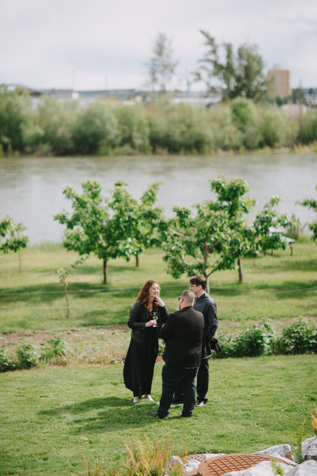 Three guests in dark clothing converse in an orchard by a river on an overcast day