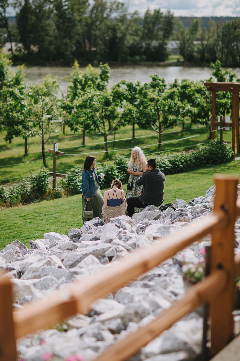 Four people gather on rocky riverbank with lush vineyards and forested hills beyond in soft daylight