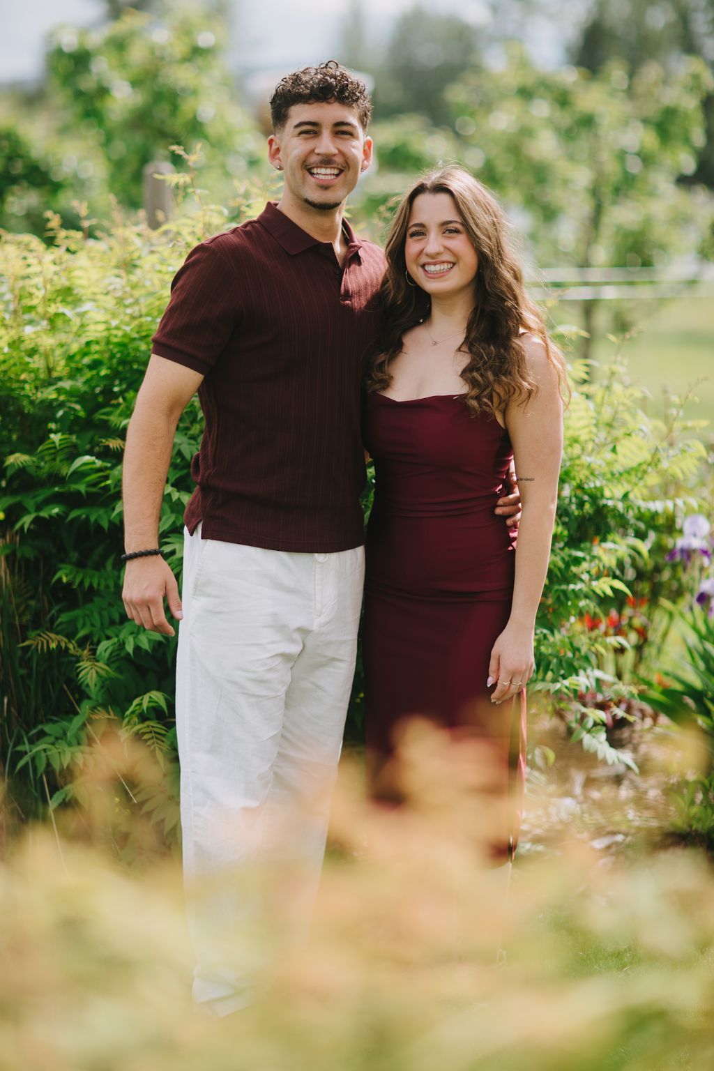 Couple in maroon and white outfits smiling outdoors with blurred green foliage background in natural daylight