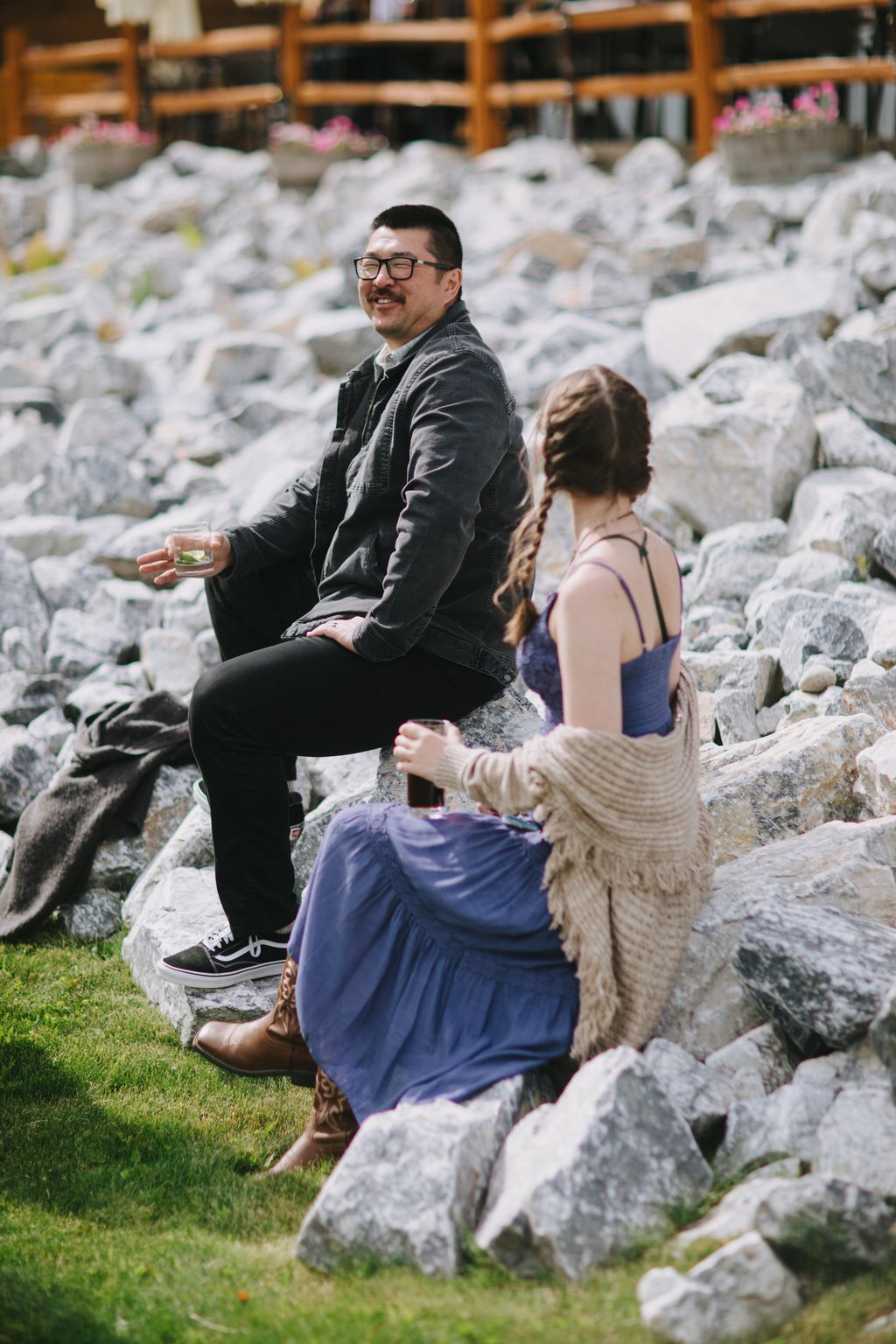 Groom in glasses and denim jacket smiles while sitting with bride on stone rocks in natural outdoor setting