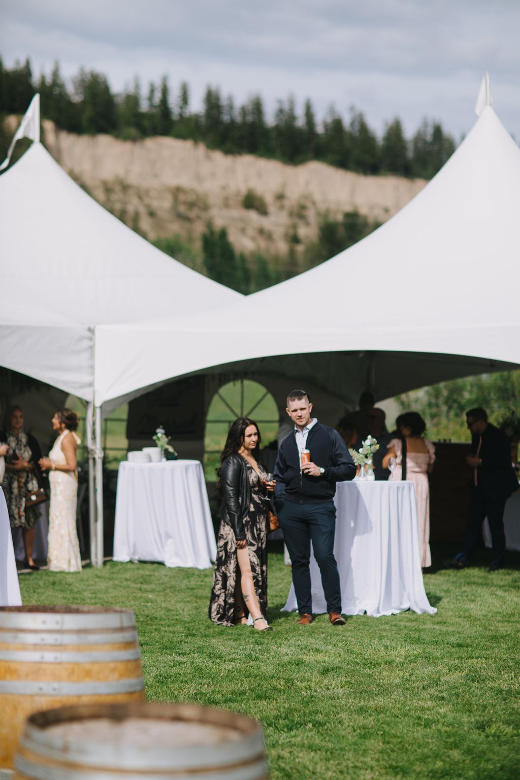 Couple stands under elegant tent with white-draped tables, holding drinks with forested hillside and overcast sky behind them
