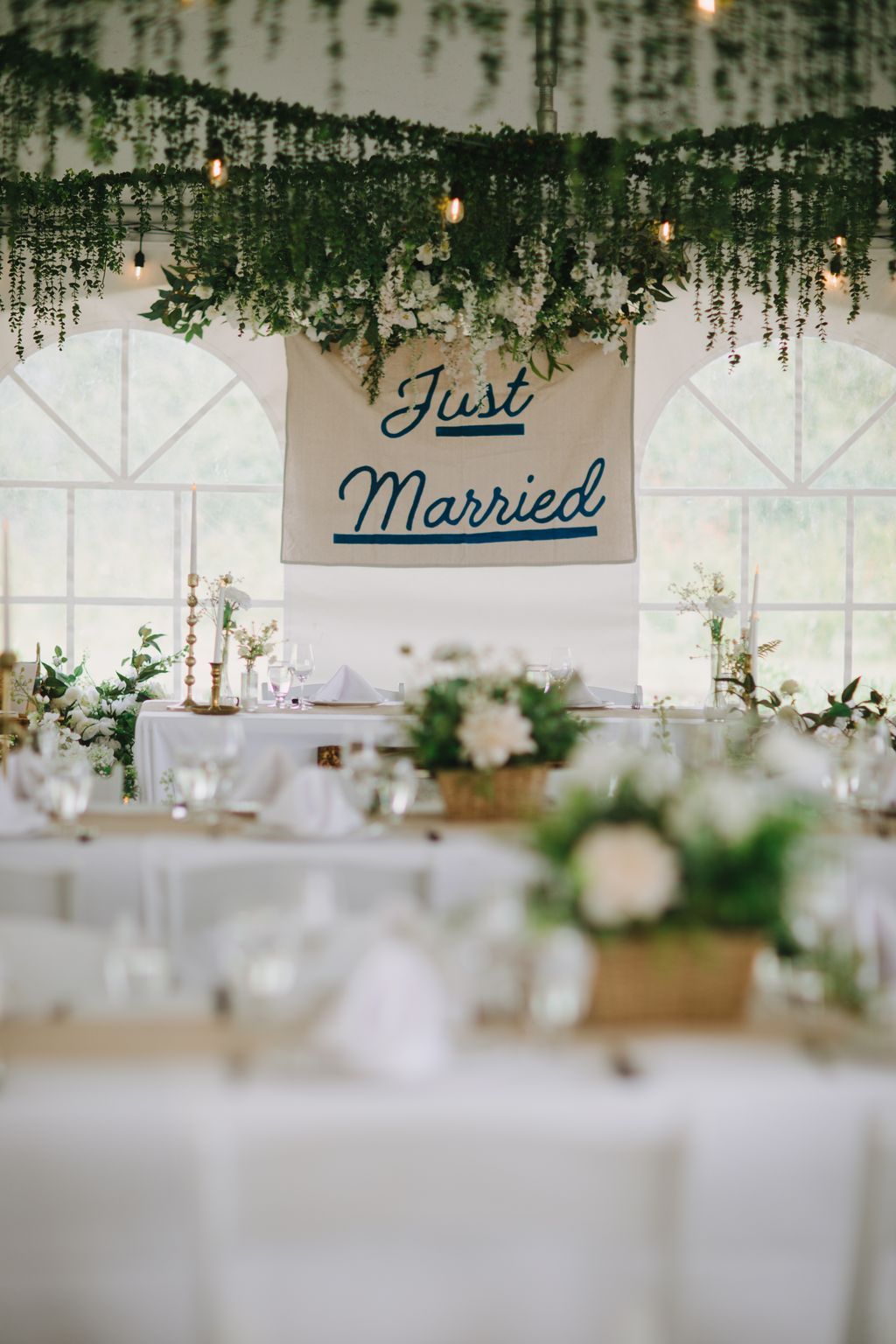 Elegant wedding reception banner reading "Just Married" in navy script, suspended above a decorated table with greenery garland, warm string lights, and whit…