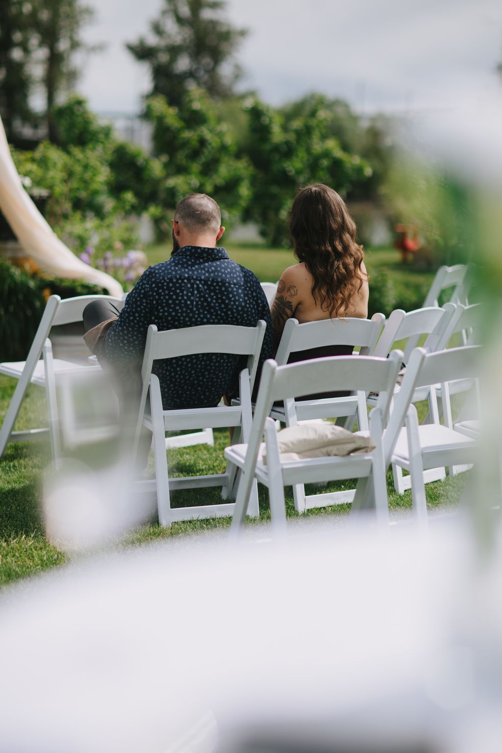 Couple seated on white chairs facing green lawn and trees during outdoor ceremony