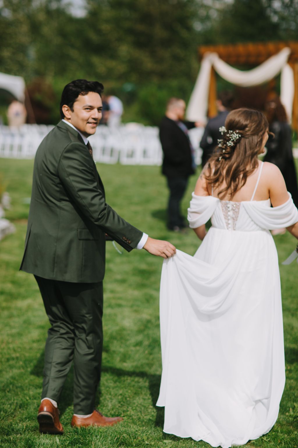 Groom in olive suit and bride in white off-shoulder dress hold hands on manicured lawn during outdoor ceremony