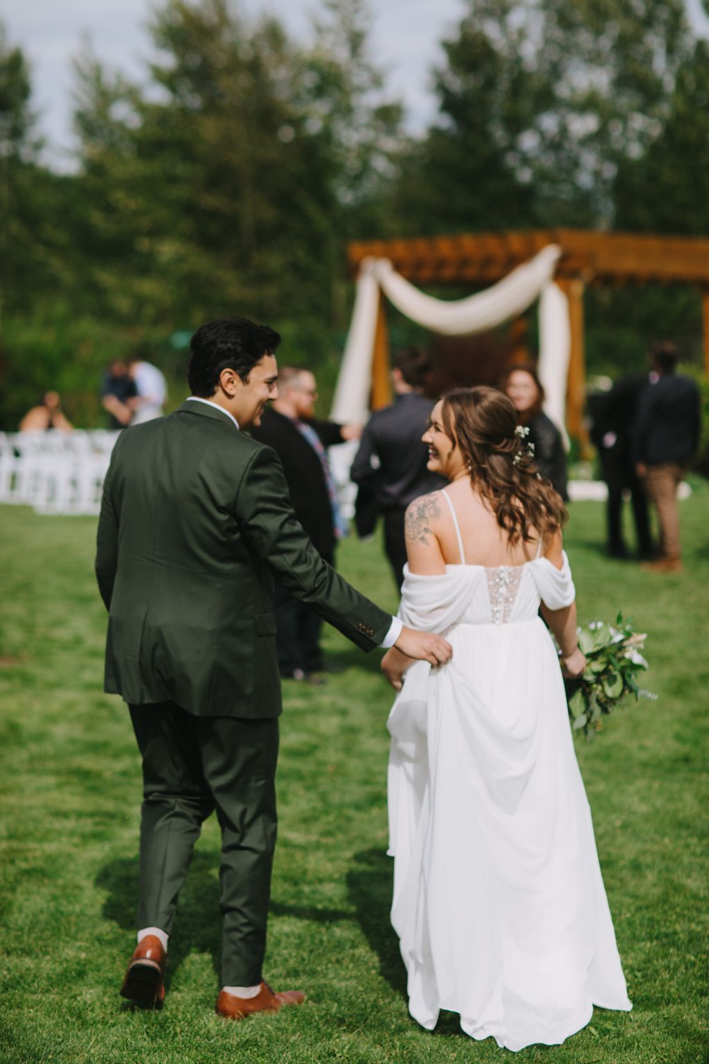 Bride and groom hold hands during outdoor ceremony with draped arch and evergreen trees in soft natural light
