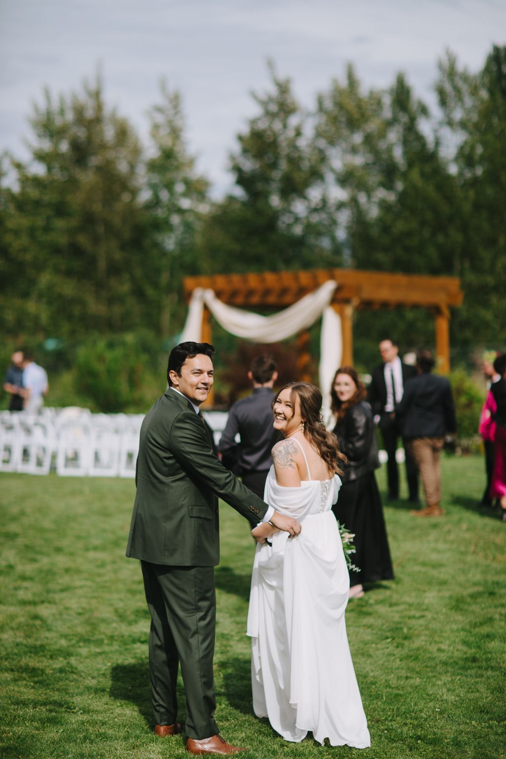 Bride in white off-shoulder dress and groom in olive suit smile at camera on grass with wooden arbor and evergreen trees behind them