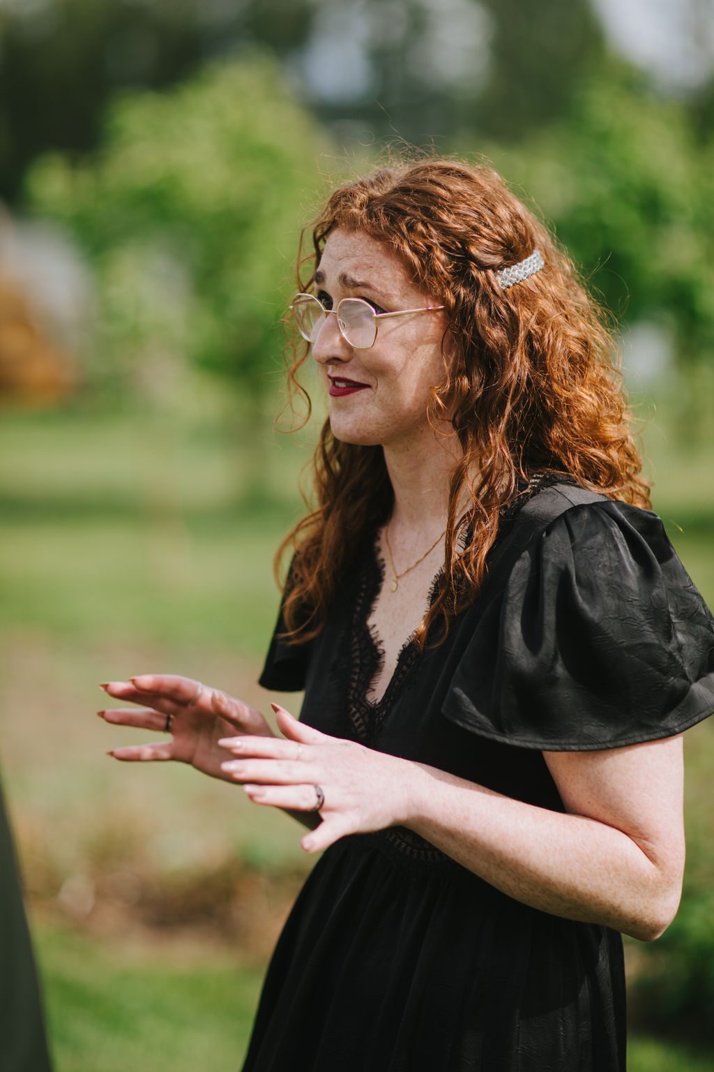 Woman with copper wavy hair and glasses smiling outdoors in natural sunlight with soft green background