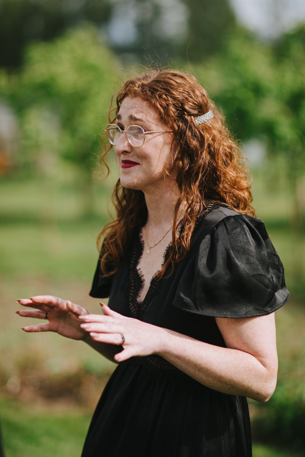 Woman with copper-red wavy hair and glasses, wearing black lace top, gestures expressively outdoors with soft green background