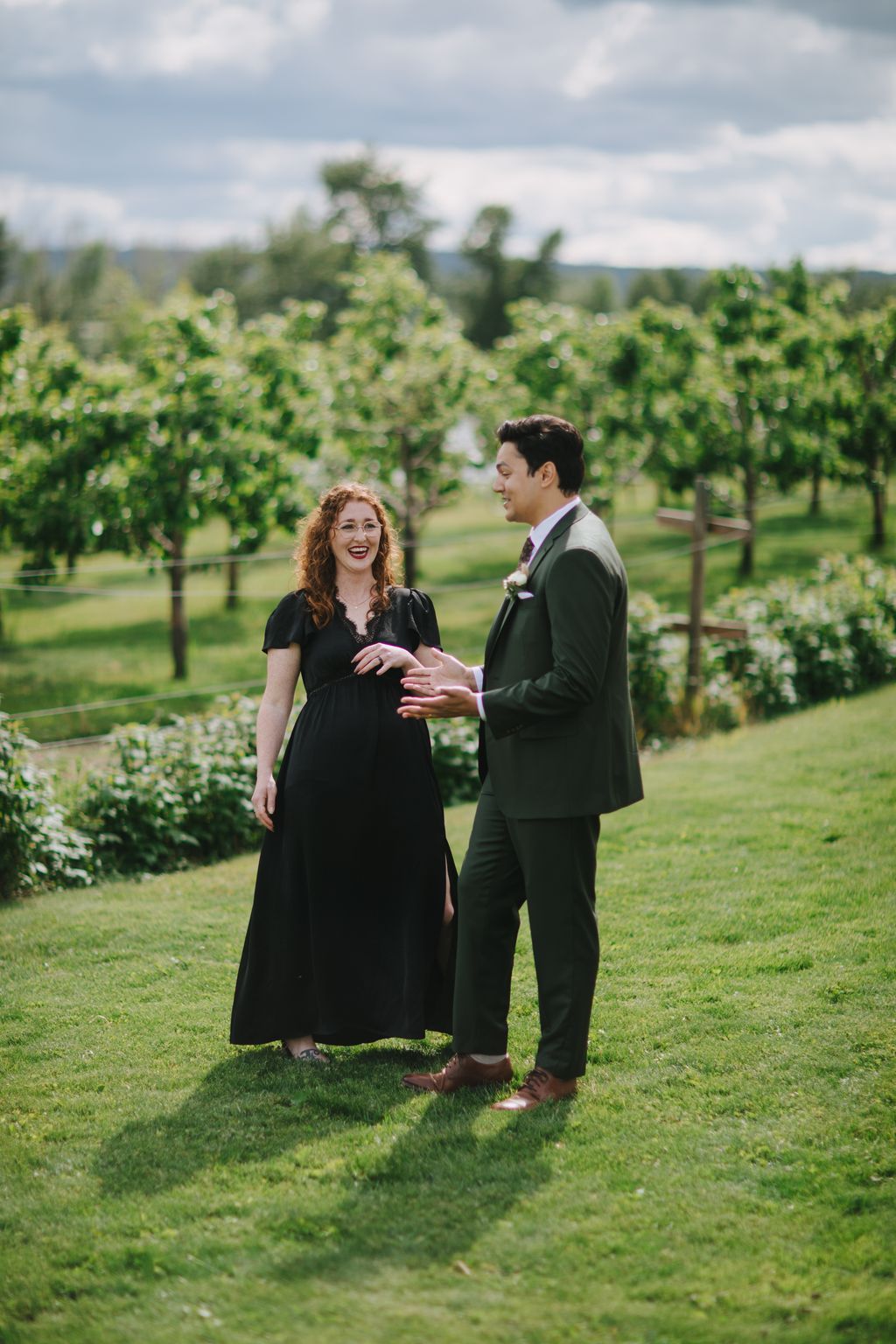 Couple holding hands in a sunlit orchard, woman in black dress with red lipstick, man in olive suit, lush green vineyard backdrop