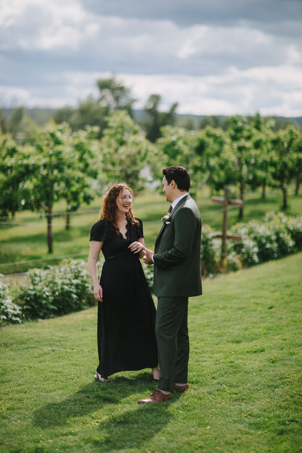 Couple in formal attire holding hands in a sunlit vineyard, smiling at each other under cloudy skies