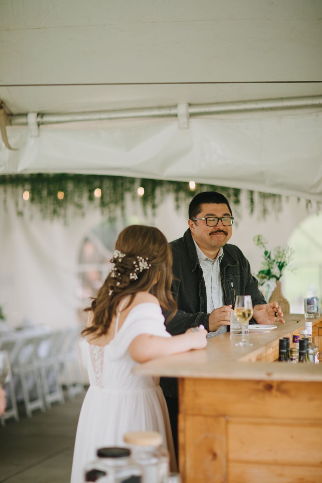 Groom in glasses and black jacket smiles at bride in white dress with pearl hair pins under tent at reception